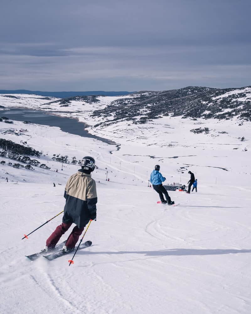 Falls Creek Ruined Caste - Skiers and snowboarders on ski run with lake in background 
