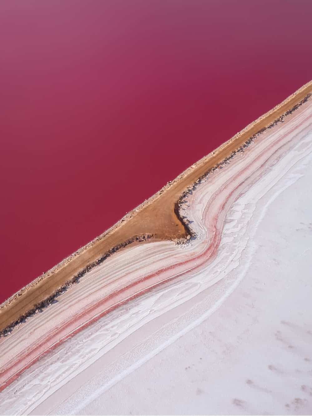 Aerial view of Hutt Lagoon's vibrant pink waters divided by a narrow earthen causeway, with salt deposits creating textured white and pink patterns along the edges.