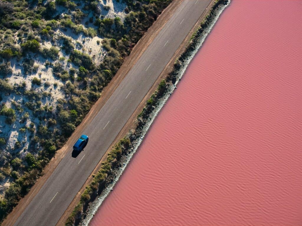 An aerial shot capturing the striking contrast between a road and the pink-hued waters of Hutt Lagoon, with a single car driving alongside this natural wonder near Port Gregory.