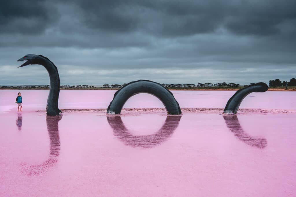 Sculptural representation of the Loch Ness Monster emerging from the pink waters of Lake Bumbunga in Australia, with a person standing nearby for scale under a dramatic cloudy sky.