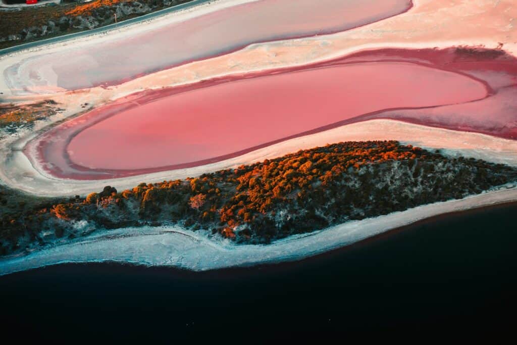 An aerial shot captures the warm glow of sunset over the pink salt lakes of Rottnest Island, contrasting with the dark blue waters of the surrounding sea and the rich greenery of the island's vegetation.